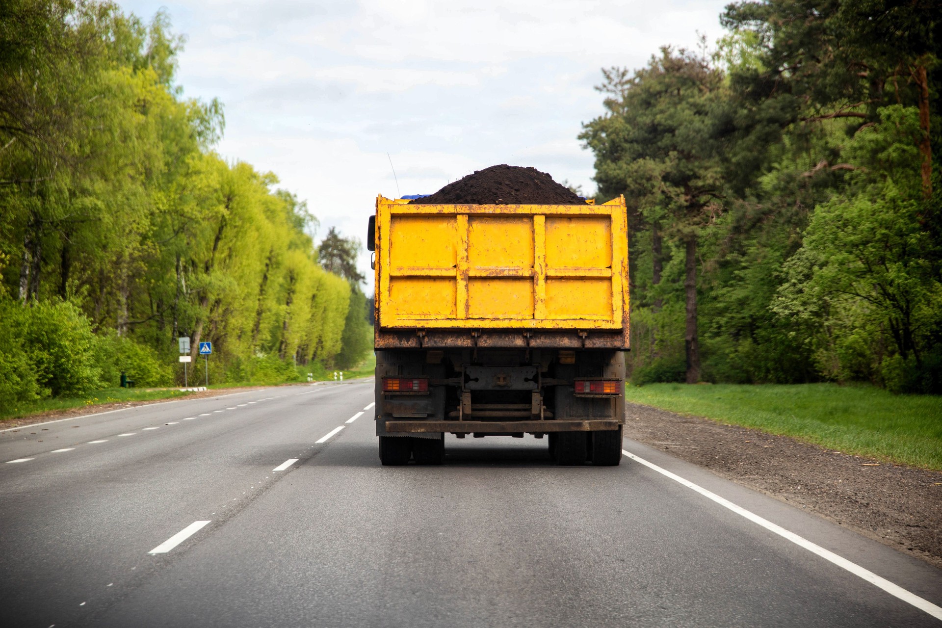A dump truck with a yellow body transports black soil along an asphalt road against the backdrop of a forest, industry